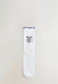 Calzino bianco a coste con un emblema ornamentale nero e grigio vicino alla parte superiore e un bordo ondulato viola, posato su una superficie bianca.