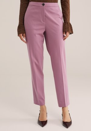 Femme portant un pantalon mauve taille haute et jambes droites, un haut en dentelle marron à manches longues, et des talons marron à bouts pointus, debout devant un fond neutre.