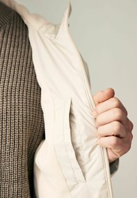 Jachetă exterioară albă, fermoarul parțial deschis, dezvăluind umplutura interioară moale și texturată de culoare bej și un pulover tricotat maro cu răscroiri subțiri în dedesubt.