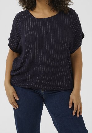 Femme portant un haut bleu marine ample à manches courtes avec de fines rayures blanches verticales et un jean bleu foncé, debout avec les mains détendues.