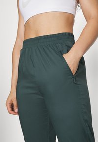 Donkergroene sportbroek met een elastische tailleband, zijzak en een gladde textuur. Kort bij de taille met een effen achtergrond.