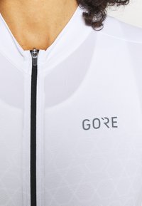 Biały rowerowy dres z czarnym zamkiem błyskawicznym i szarym logo „GORE”, z teksturowanym wzorem na tkaninie i dopasowanym krojem.