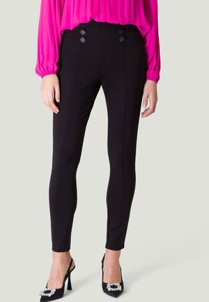 Femme portant un pantalon noir taille haute avec des boutons décoratifs, associé à des talons noirs avec boucles en strass, debout devant un fond uni.
