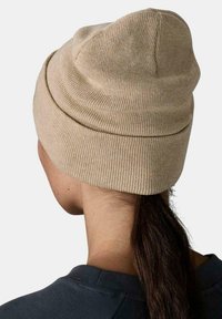 Gorro beige de punto llevado en la parte trasera de la cabeza, cubriendo las orejas, con cabello largo y oscuro salido por la parte trasera.