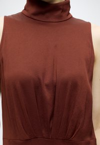 Personne portant un haut marron sans manches à col roulé avec des détails plissés sous la poitrine, montrée des épaules à la taille.