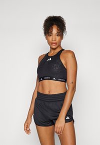 Črn športni crop top z teksturiranim vzorcem, logotip na pasu; v kombinaciji s črnimi kratkimi hlačami z elastičnim pasom in logotipom.