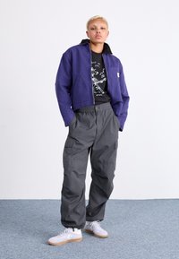 Lila cropped jacka, svart grafisk t-shirt, grå cargobyxor, vita sneakers med gummisålar. Enkelt design, standard passform, funktionella fickor.