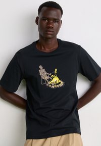 Camiseta de algodón negra con un gráfico de un esqueleto sentado junto a una fogata, con llamas amarillas y la palabra "Peligro" en una fuente negrita y divertida.