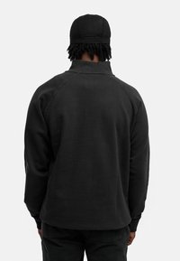 Schwarzer Pullover mit hohem Kragen und Raglanärmeln. Weicher Stoff, lockere Passform und minimale Nähte. Außerdem enthält er eine passende Kappe.