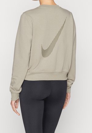 Osoba ubrana w beżową bluzę z dużym pasiastym logo Nike na plecach oraz czarne dopasowane legginsy, pokazana od tyłu.