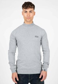 Maglione grigio, colletto alto, design aderente, polsini e orlo a coste, presenta il logo "WRONG FRIENDS" sul lato sinistro del petto. Indossato con jeans scuri.