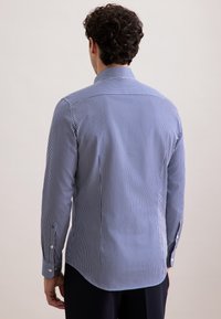 Uomo di spalle che indossa una camicia a maniche lunghe a righe verticali blu e bianche e pantaloni scuri su uno sfondo neutro.