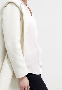 Manteau couleur crème avec une capuche, texture en laine, associé à un chemisier rose pâle. La tenue présente une coupe ajustée et un devant avec des boutons.