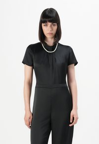 Young woman with straight dark hair wearing a black short-sleeve jumpsuit and a single strand pearl necklace, facing forward.
