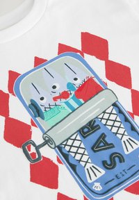 Bílý tričko s barevným grafikou plechovky, na které jsou štětce, nůžky a červený předmět. Pozadí má červené diamantové vzory.