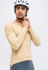 Νέος άνδρας με μπεζ ποδηλατική φανέλα που δένει το λουρί ενός λευκού κράνους ποδηλάτου, στέκεται μπροστά σε απλό φόντο.