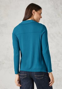 Türkisfarbener Langarmshirt mit Kragen, ausgestattet mit einer Rückenpasse, seitlichen Schlitzen und glatter Textur, kombiniert mit dunkelblauen Jeans.