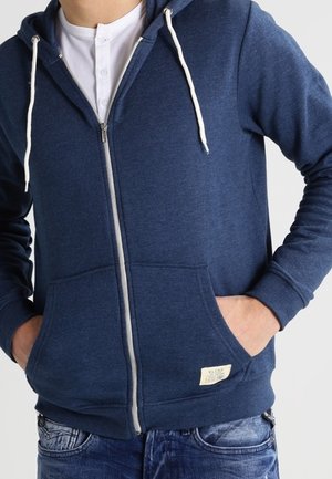 Persona che indossa un felpa con zip blu navy con lacci bianchi, una maglietta henley bianca sotto e jeans blu strappati, con le mani nelle tasche della felpa.