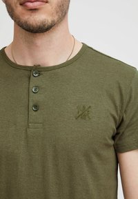 Olivgrünes Baumwoll-T-Shirt mit Rundhalsausschnitt, das drei Knöpfe und ein dezentes gesticktes Logo-Detail auf der linken Brust hat.