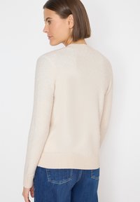 Suéter de punto beige con una textura suave, dobladillo acanalado y escote redondo. Llevado con jeans azules, visto desde la parte de atrás.