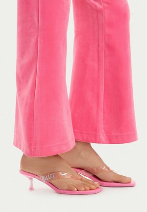 Füße, die pinkfarbene Sandalen mit Absatz, durchsichtigen Riemen und Strass-"Juicy"-Detail tragen, kombiniert mit pinken samtweiten Hosen.