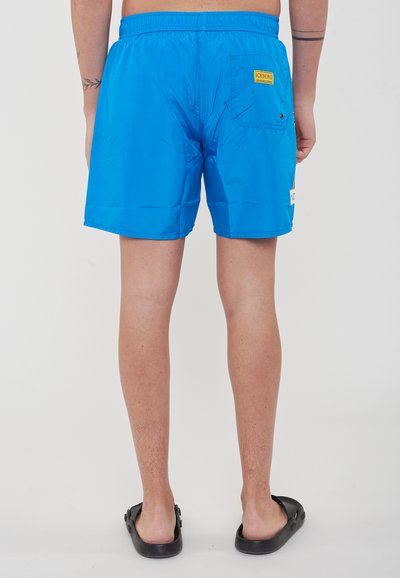 Blaue Badehorts mit elastischem Bund, einer Gesäßtasche mit gelbem Markenetikett und einer glatten, leichten Textur.