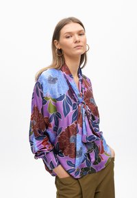 Blusa di seta con un vivace motivo floreale in tonalità di blu, rosso e verde, con un dettaglio a fiocco sul collo e maniche ampie.