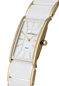 Reloj de cerámica blanca con una esfera rectangular, acentos en tono dorado, marcadores de hora dorados y un mecanismo de cuerda coronado.