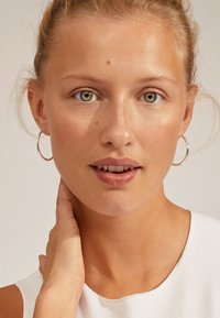 Modelo con una blusa sin mangas blanca, luciendo pendientes de aro. La piel tiene una textura suave; sus rasgos incluyen ojos verdosos y maquillaje natural.