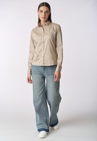Camicia lunga beige chiaro con colletto, chiusura a bottoni e ricamo sottile, abbinata a jeans larghi blu e scarpe bianche.