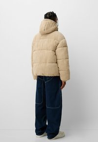 Corduroy puffer jacket in light beige with a hood, featuring a quilted design. Paired with dark blue wide-leg jeans and white sneakers.