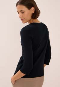 Vrouw met kort bruin haar in een zwart shirt met lange mouwen en beige broek, met handen in de zakken, terwijl ze over haar schouder kijkt tegen een effen achtergrond.