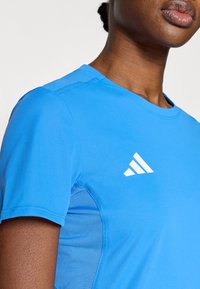 Modra športna srajca s kratkimi rokavi, ki ima teksturirano ploščo na strani in belo značko na prsih. Gladek, tkanina, ki odvaja vlago.