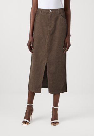 Vrouw draagt een bruine midirok met een split aan de voorkant, gecombineerd met witte sandalen met hoge hakken en een wit topje, staand tegen een neutrale achtergrond.