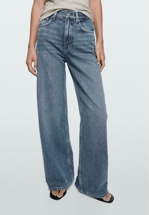 Person iført højttaljede, viddebenede blå jeans med forlommer og knaplukning, kombineret med sorte åbne sandaler på en ensfarvet baggrund.