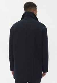Cappotto di lana blu navy con colletto alto e spalle strutturate, caratterizzato da un design pulito e minimalista e da una vestibilità su misura.