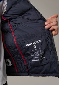 Navy puffer jacket interior with red accents, featuring a label for Strellson, symbols for recycled filling, and a small side pocket.