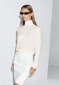 Pullover col roulé beige à manches longues associé à une jupe blanche ajustée. Modèle portant des lunettes de soleil rectangulaires foncées devant un fond gris.