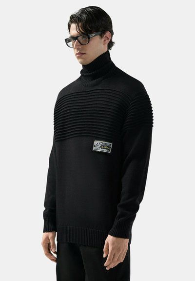 Uomo che indossa un maglione a coste nero con collo alto e una toppa sul petto, pantaloni neri e occhiali rettangolari neri, in piedi davanti a uno sfondo bianco.