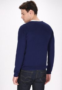 Temno modra jopica z dolgimi rokavi, krožnim izrezom in gladko teksturo. Nosa z temnimi jeans hlačami z vidnimi zadnjimi žepki in šivi.
