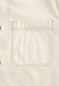 Tela de camisa en un tono beige claro con textura acanalada en diagonal, que presenta un bolsillo cuadrado en el pecho delineado con costuras contrastantes y botones de metal plateado.