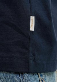 Κοντινό πλάνο σε μια ετικέτα ρούχου από ύφασμα ναυτικού μπλε με το κείμενο "Originals" ραμμένο στη ραφή πάνω από μπλε τζιν παντελόνια.