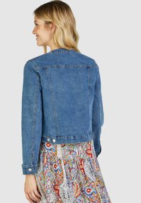 Blaue Jeansjacke mit tailliertem Schnitt, mit Knopfdetails an den Ärmeln und am unteren Saum, von hinten auf einem gemusterten Rock gezeigt.