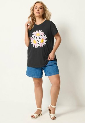 Kobieta ubrana w czarny t-shirt z różowo-białym nadrukiem stokrotki, niebieskie denimowe szorty i białe sandały, stojąca na jednolitym tle.