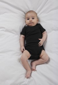 Baby ligger på hvide lagner iført en sort heldragt, kigger op med et neutralt udtryk og let sammenknebne læber.