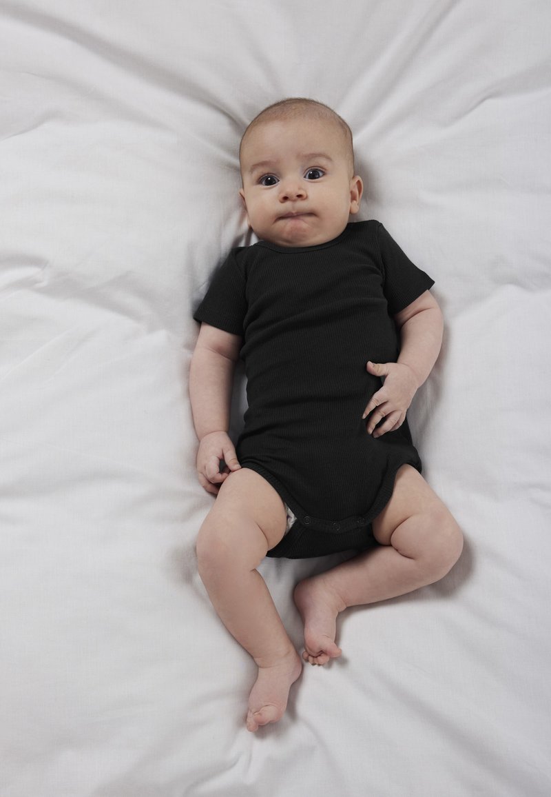 Baby ligger på hvide lagner iført en sort heldragt, kigger op med et neutralt udtryk og let sammenknebne læber.