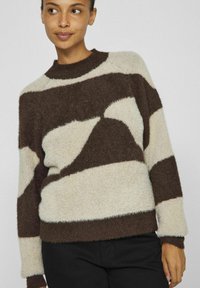 Braun- und cremefarbener gemusterter Pullover mit einer fuzzy Textur, oversized Schnitt und geripptem Ausschnitt, kombiniert mit schwarzen Hosen.
