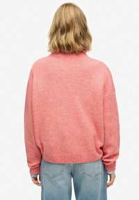 Vrouw met schouderlang blond haar, gekleed in een losse roze geribbelde trui en lichtblauwe jeans, van achteren gezien tegen een witte achtergrond.