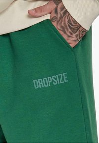 Grüne Sweatpants mit einer weichen Textur, die eine Seitentasche und den Aufdruck "DROPSIZE" in Hellgrau über den Stoff verfügen.