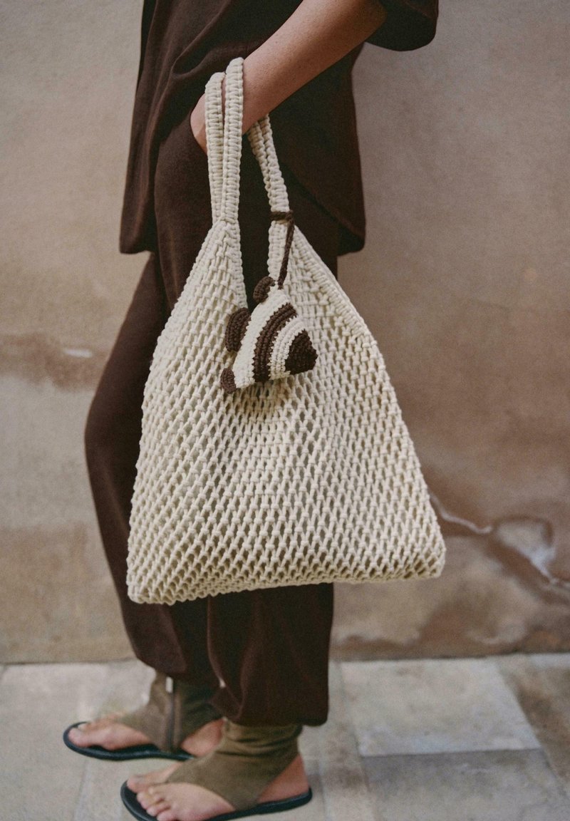 Personne vêtue de vêtements sombres tenant un grand sac fourre-tout tricoté couleur crème avec un petit ornement tricoté marron et crème attaché.